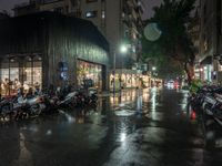 a bunch of motorcycles parked on the street at night on a rainy sidewalk in a busy city