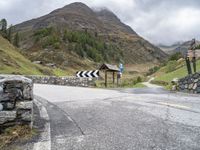 a winding mountain side road with a sign on the left and a stone wall on the right