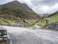 a winding mountain side road with a sign on the left and a stone wall on the right