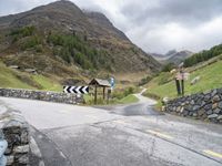 a winding mountain side road with a sign on the left and a stone wall on the right