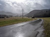 a wet road with telephone lines and a field behind it in the rain and storm clouds