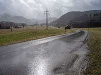 a wet road with telephone lines and a field behind it in the rain and storm clouds