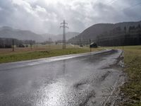 a wet road with telephone lines and a field behind it in the rain and storm clouds