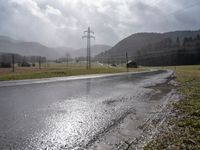 a wet road with telephone lines and a field behind it in the rain and storm clouds