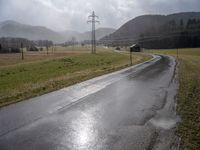 a wet road with telephone lines and a field behind it in the rain and storm clouds