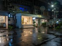 a wet road with a small store next to it and the neon light above the buildings