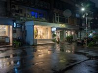 a wet road with a small store next to it and the neon light above the buildings