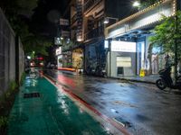 a wet street at night with a motorcycle parked on the sidewalk beside it and a building near by