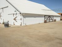 a photo of a white barn that is outside of it's yard, and there is an open space with barrels