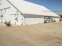 a photo of a white barn that is outside of it's yard, and there is an open space with barrels