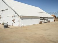 a photo of a white barn that is outside of it's yard, and there is an open space with barrels