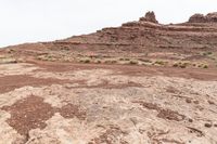 dirt road in desert near rock formations with sparse grass and shrubs around it under a white cloudy sky