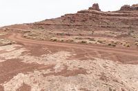 dirt road in desert near rock formations with sparse grass and shrubs around it under a white cloudy sky