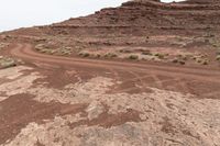 dirt road in desert near rock formations with sparse grass and shrubs around it under a white cloudy sky