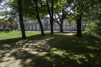 large white house with several trees in a grassy area near it and another building in the distance