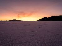 a very wide open field covered with white snow and mountains behind it at sunset time