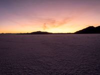 a very wide open field covered with white snow and mountains behind it at sunset time