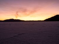 a very wide open field covered with white snow and mountains behind it at sunset time