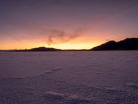 a very wide open field covered with white snow and mountains behind it at sunset time