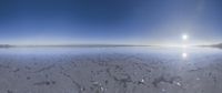 a wide view of the sun in a cloudy sky over the ocean with a snow covered shoreline