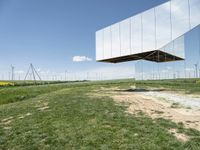 a large mirror structure in a field with wind turbines in the background behind it and a blue sky above