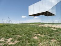 a large mirror structure in a field with wind turbines in the background behind it and a blue sky above