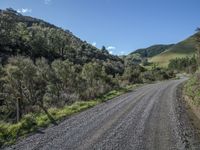 the dirt road is going into the mountains and forests on one side of the road, a hill in the other