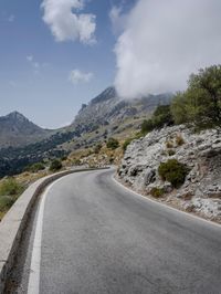 a road that is winding in front of mountains under a cloudy sky over some vegetation