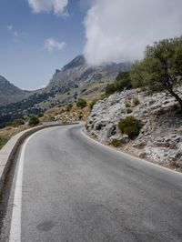 a road that is winding in front of mountains under a cloudy sky over some vegetation