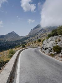 a road that is winding in front of mountains under a cloudy sky over some vegetation