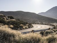 an empty winding road with no cars on it near a hill side with grassy bushes