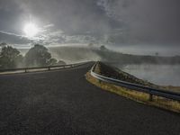 the view of a winding road going over a body of water with mist and clouds