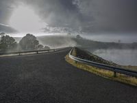 the view of a winding road going over a body of water with mist and clouds