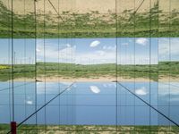 large glass cube reflecting water and grass with mountains in the distance near it, on the floor of square open area