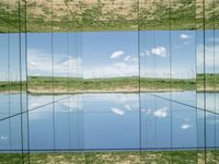 large glass cube reflecting water and grass with mountains in the distance near it, on the floor of square open area