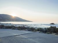a parking lot with sand and hills in the background and the sun setting over a beach