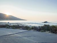 a parking lot with sand and hills in the background and the sun setting over a beach
