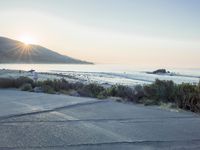 a parking lot with sand and hills in the background and the sun setting over a beach