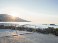 a parking lot with sand and hills in the background and the sun setting over a beach