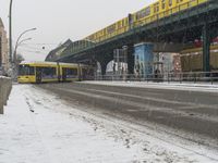 snow is falling onto the road and the yellow commuter train is crossing over the bridge