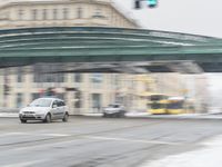 cars going down a city road with an overpass behind them covered in snow and cars passing on the street