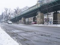 a snow covered street with traffic next to bridge and building overpasss on the left