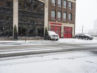 two cars driving through the snow on a snowy day in front of an old brick building