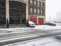 two cars driving through the snow on a snowy day in front of an old brick building