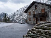 a car in front of a house with some logs piled near it and snow on the mountains behind