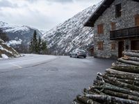 a car in front of a house with some logs piled near it and snow on the mountains behind