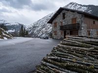 a car in front of a house with some logs piled near it and snow on the mountains behind