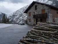 a car in front of a house with some logs piled near it and snow on the mountains behind