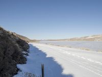 a small dirt road leads to a body of water and hills with snow all around