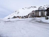 there are cars on the snow road near the buildings of a resort and mountains in the background